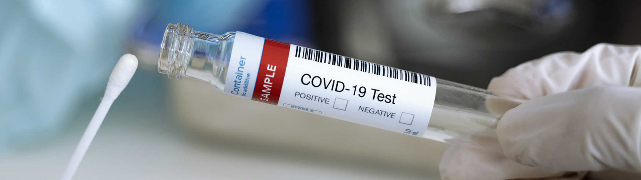 A person in gloves holds up a q-tip to a vial labeled "COVID-19 Test"