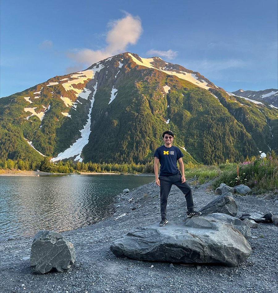Nick Tran stands on the beach in front of a mountain wearing a University of Michigan Industrial and Operations Engineering t-shirt.