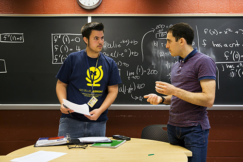 Two students discuss a problem in front of a black board with equations.