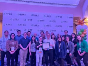 Many people stand in front of INFORMS banner holding an award from HFES