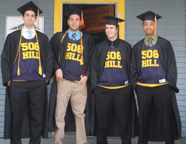 Four people stand on a porch wearing "508 Hill" shirts while in graduation cap and gowns