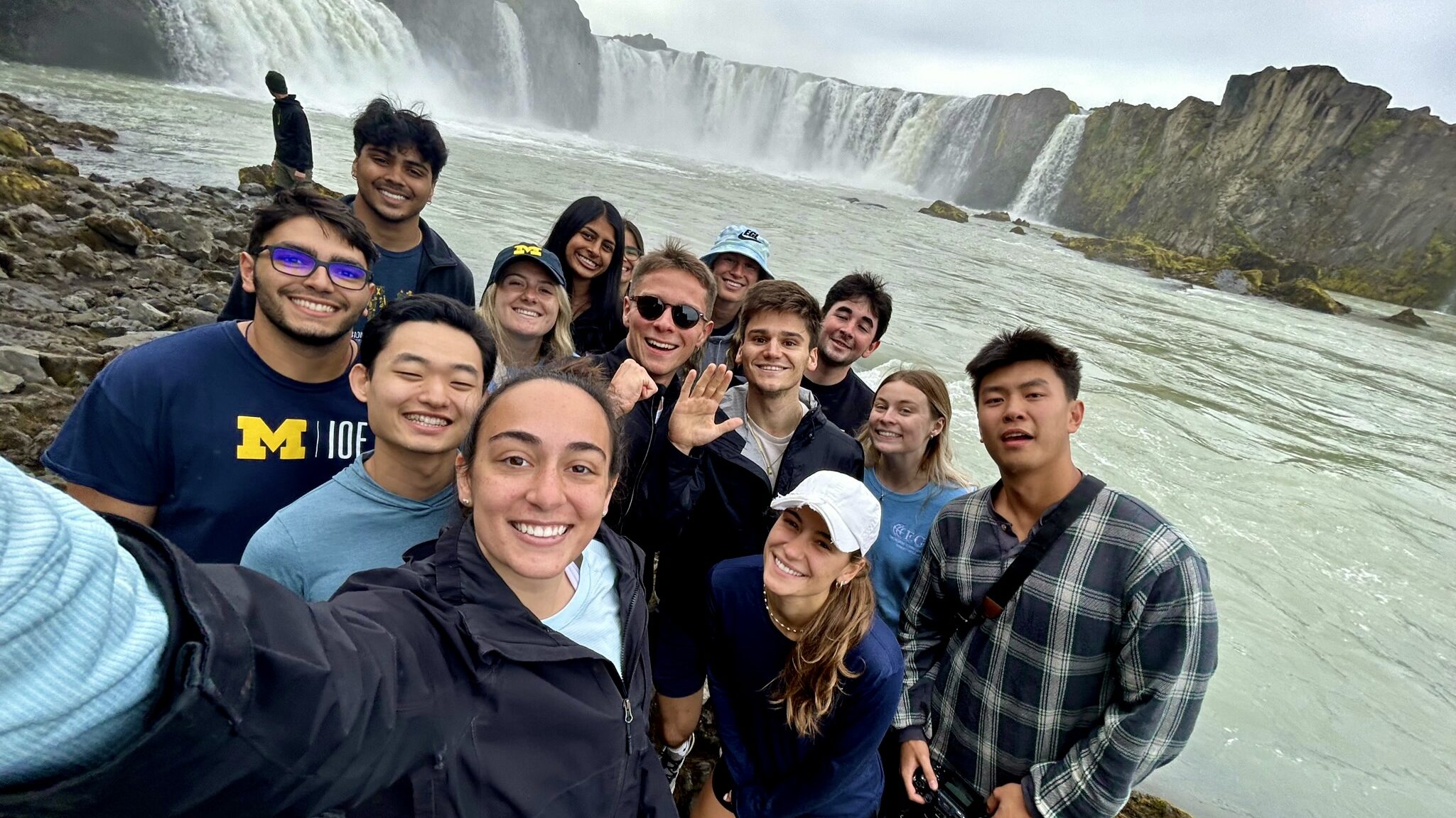A student takes a selfie of herself and 13 other people in front of a large waterfall and pool of water.