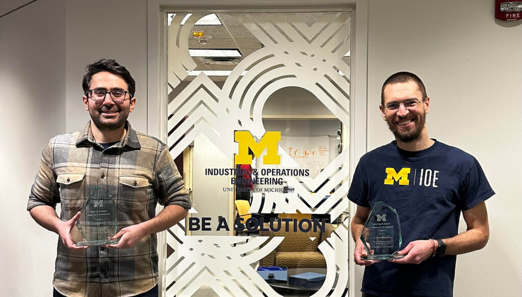 Two men hold glass awards in front of a window with a sticker that reads "Industrial and Operations Engineering University of Michigan, Be a solution"