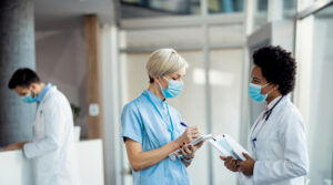 Women in a hospital setting wearing lab equipment and masks talk to each other while taking notes