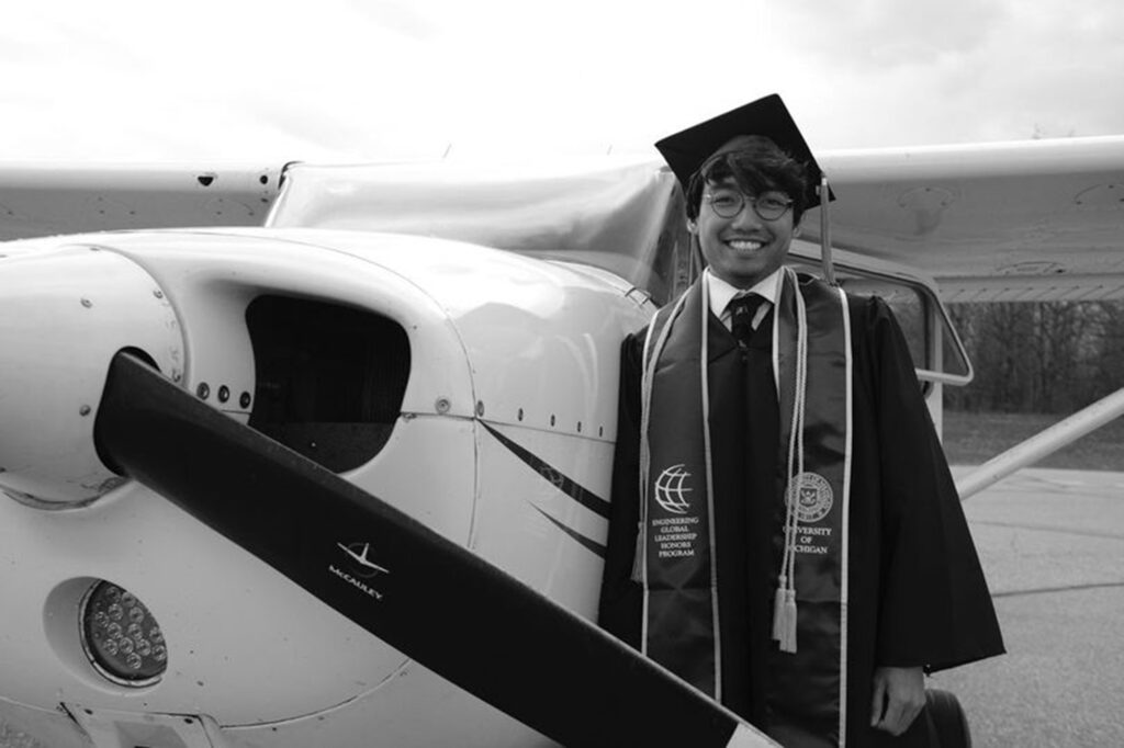 Nick Tran stands in his graduation clothes next to a small airplane in a hanger
