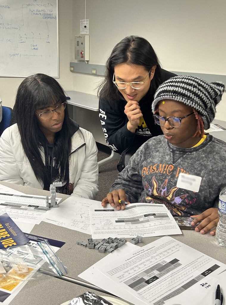One man with long black hair and glasses helps two high school students with a math problem while seated at a table. 