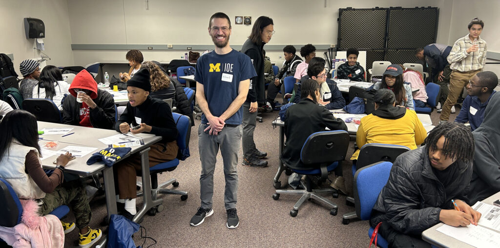 A man smiles in a crowded room surrounded by high school students sitting at tables working on math problems.