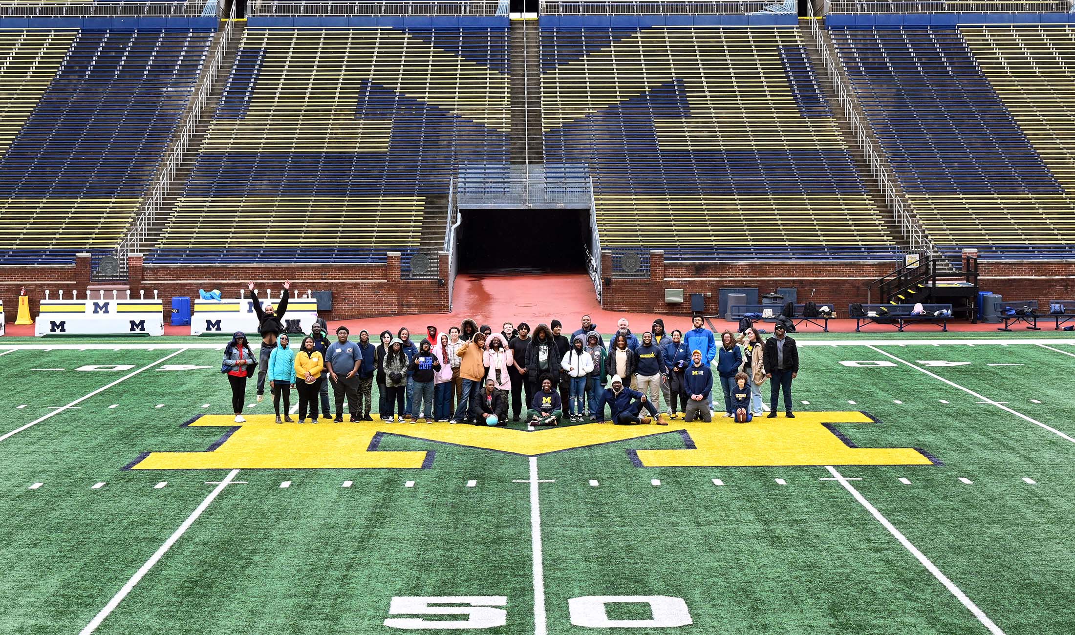 A large group of people take a photo standing on the Michigan M at the 50 yard line in Michigan stadium.