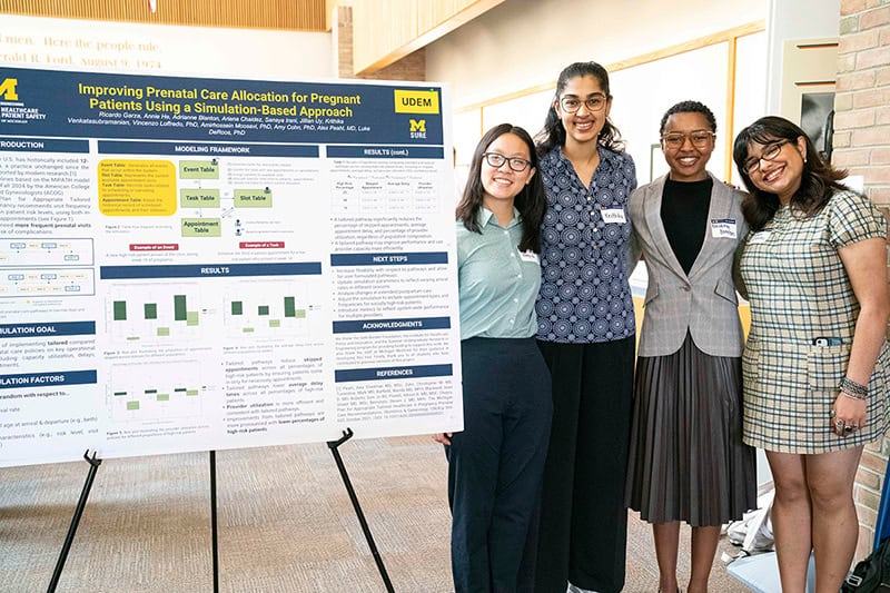 Four women stand to the right of a research poster titled "Improving Prenatal Care Allocation for Pregnant Patients Using a Simulation-Based Approach"