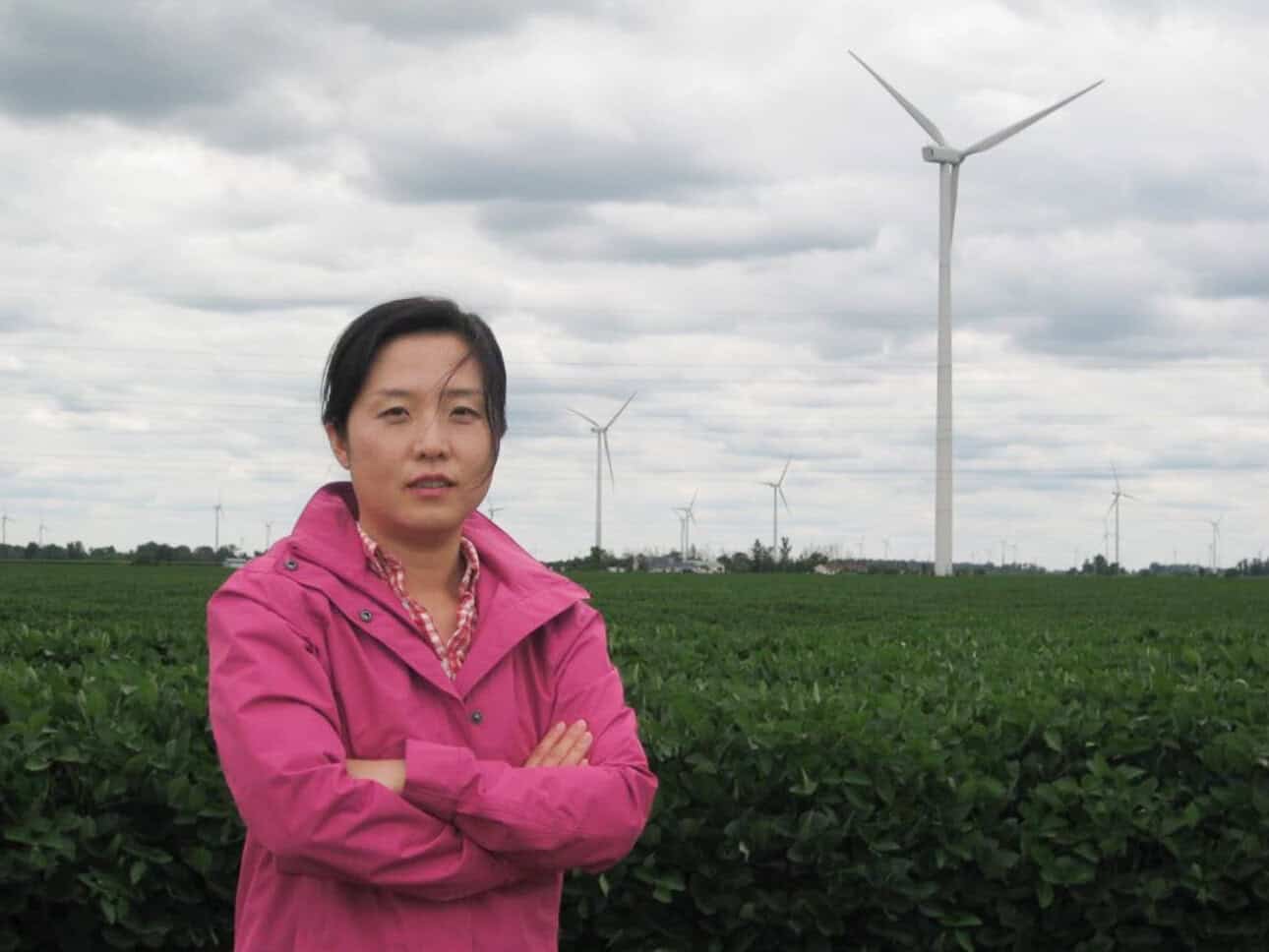 Eunshin Byon stands with arms crossed in front of a field containing several large metal wind turbines