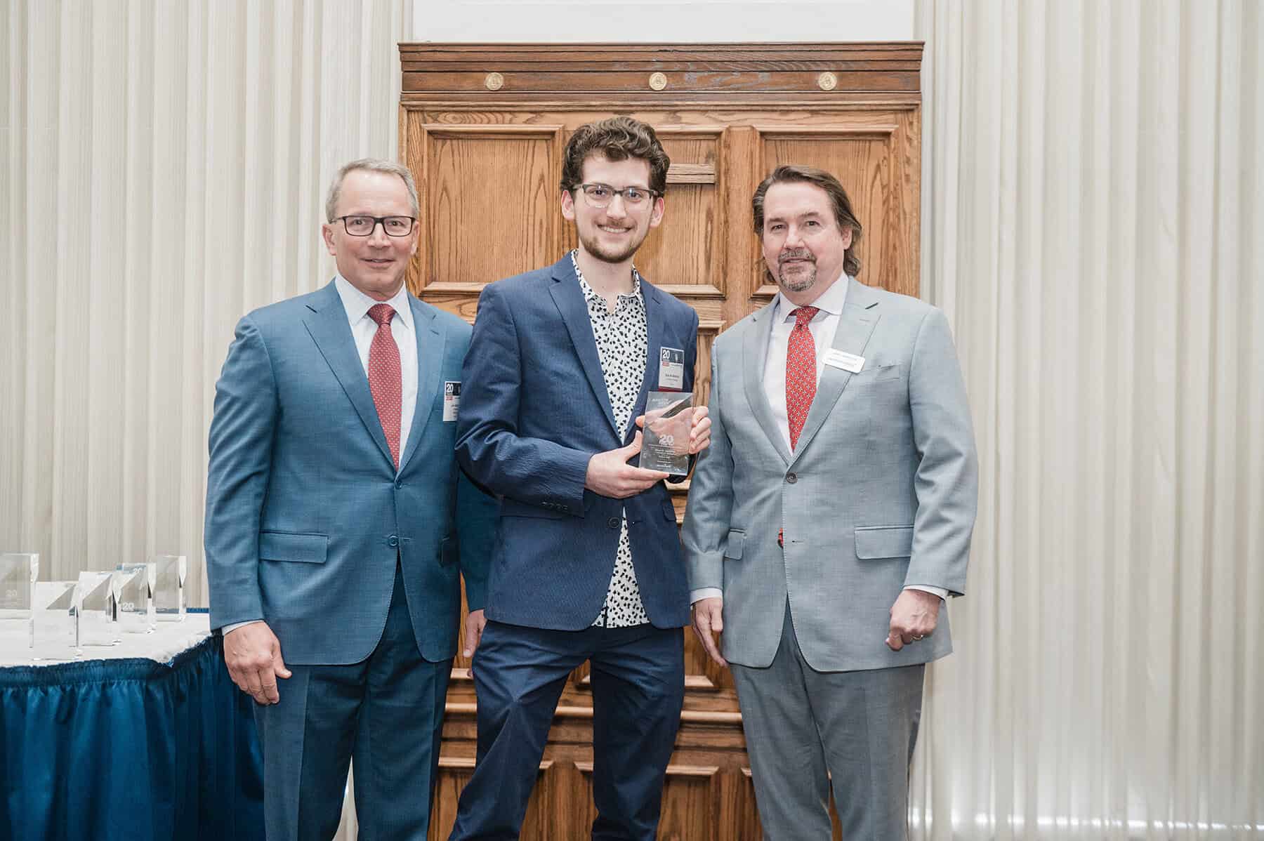 Sinan Abdulhak accepts a glass award while two men stand either side of him. They all wear formal attire.