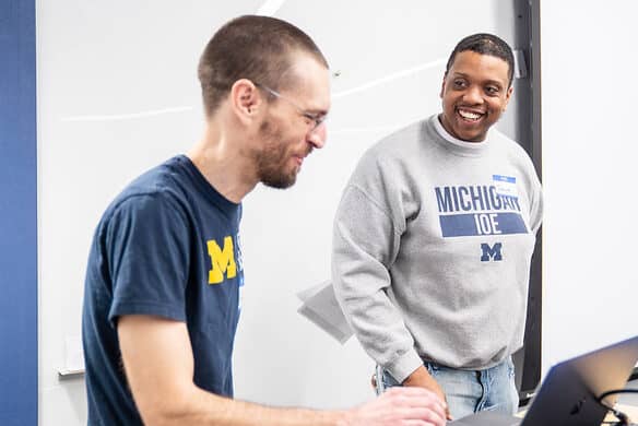 A person smiles while looking toward another person on a computer