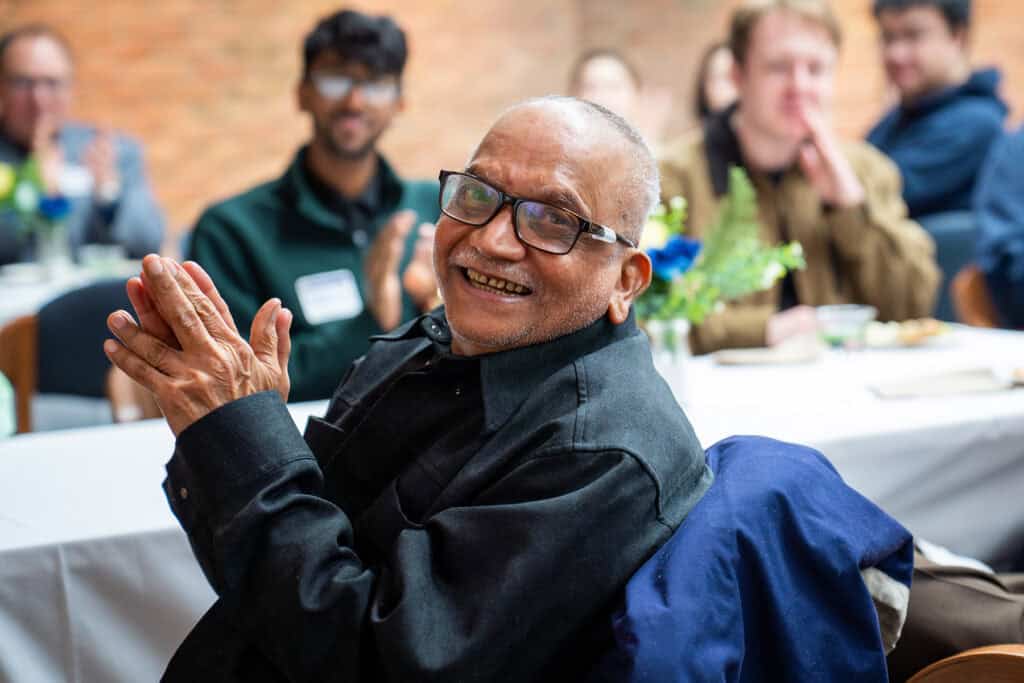 Katta Murty man wearing glasses and a navy button down claps while sitting in a crowded room.