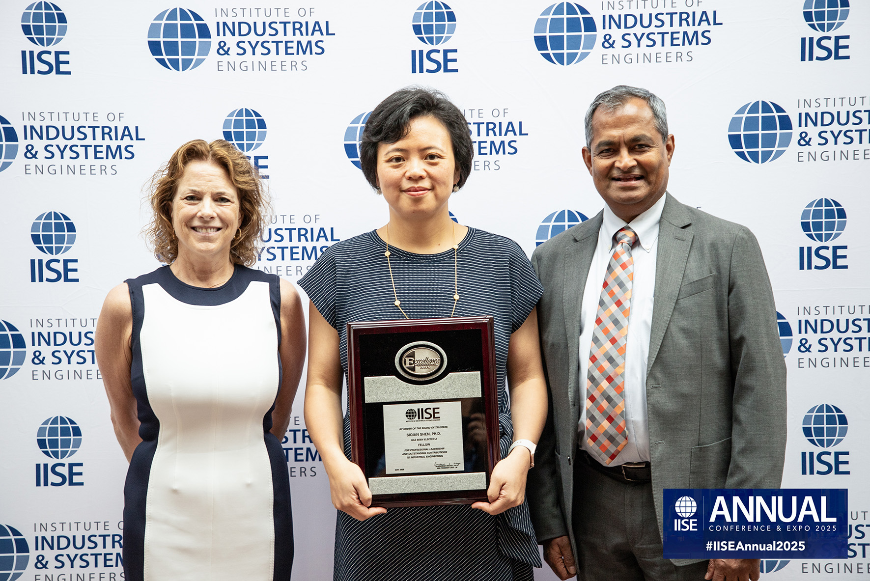 Three people stand in front of photo backdrop, middle person holding an award plaque.