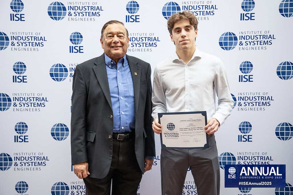 Two people stand in front of photo backdrop, one holding paper award.